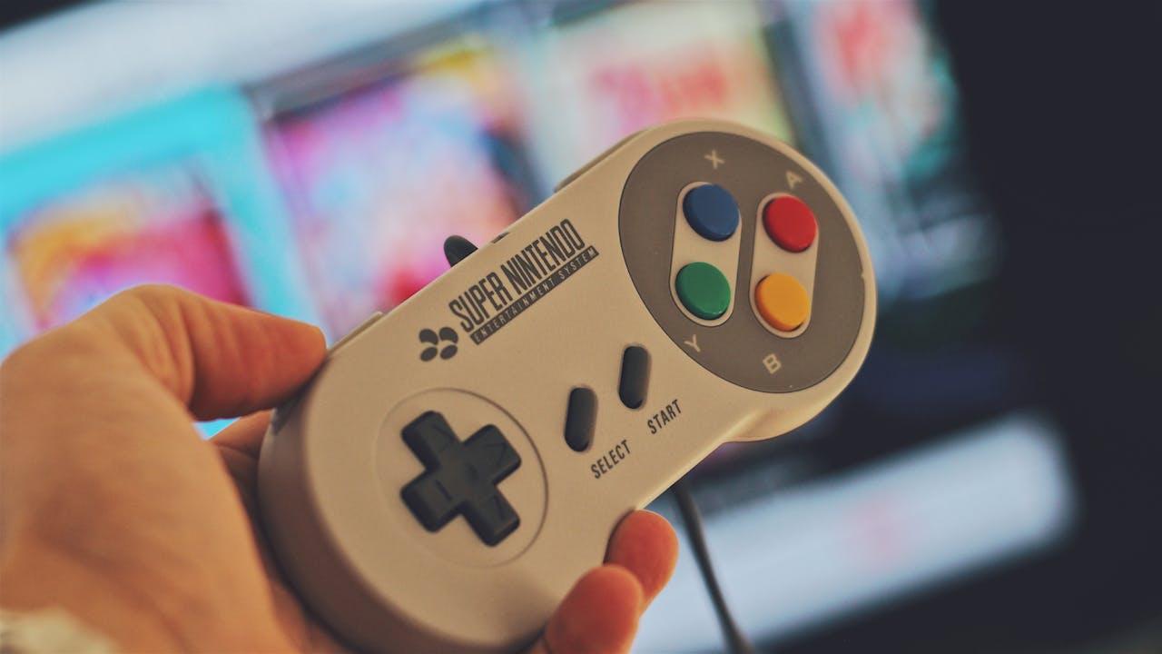 Home Hand holding a classic Super Nintendo game controller indoors with a blurred screen in the background.